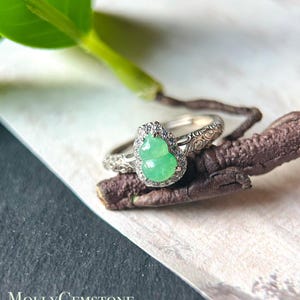Könnte beinhalten: Ein silberner Ring mit einem birnenförmigen, hellgrünen Edelstein, umgeben von kleinen, klaren Steinen. Der Ring hat ein verziertes, graviertes Design. Das Bild zeigt auch ein grünes Blatt und den Text "MollyGemstone".