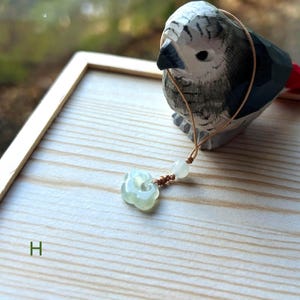 Puede incluir: Un colgante de jade verde claro con forma de flor, colgado de un cordón marrón con una pequeña perla blanca. El colgante está sobre una superficie de madera con la letra "H" visible en la esquina inferior izquierda.