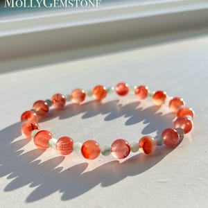 May include: A bracelet made of orange and white gemstones, with pale green beads between them. The round beads are displayed on a white surface. The text "Molly Gemstone" is visible in the upper left corner.