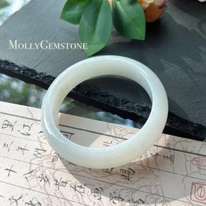 Può includere: Un braccialetto bangle in giada verde pallido. Il braccialetto circolare e lucido è appoggiato su un foglio di carta con caratteri asiatici. Il testo "MOLLY GEMSTONE" è visibile. Un gioiello classico.