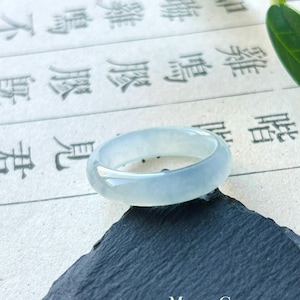 Pode incluir: Um anel de jade verde pálido translúcido. O anel é liso e polido, com uma banda arredondada. Está sobre uma superfície escura e texturizada, com um fundo de escrita manuscrita. O texto "MollyGemstone" é visível no canto inferior direito.