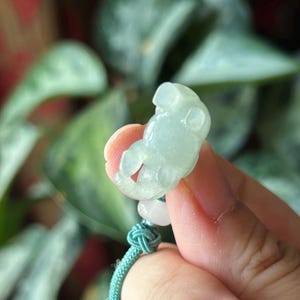 May include: A light green jade carving of a small bear. The bear is held by a hand, with a teal cord and a white bead attached. The background is blurred green foliage.