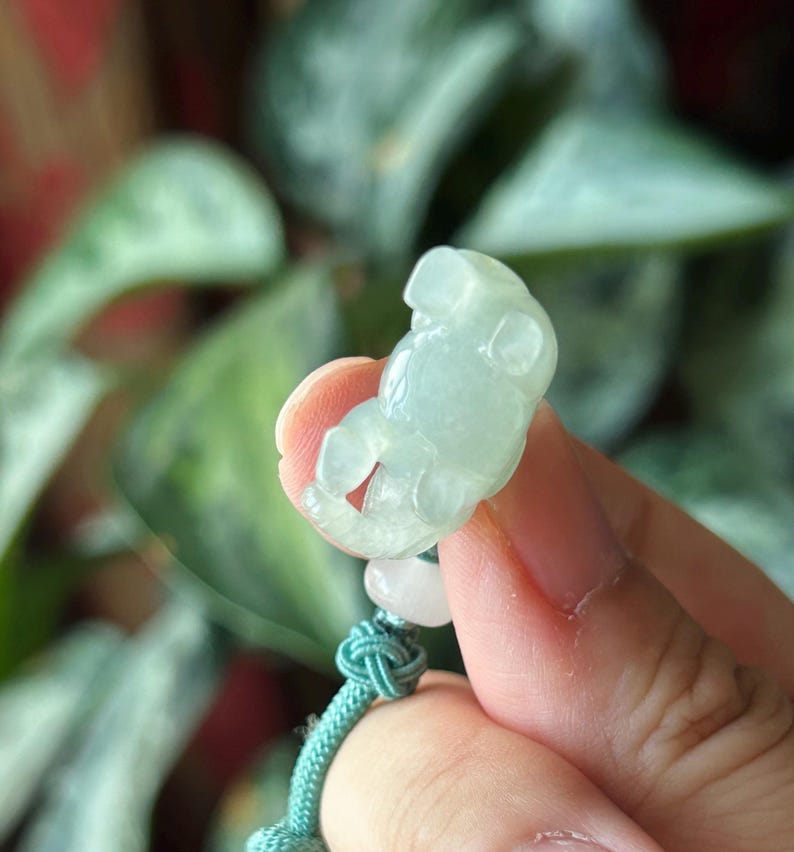 May include: A light green jade carving of a small bear. The bear is held by a hand, with a teal cord and a white bead attached. The background is blurred green foliage.