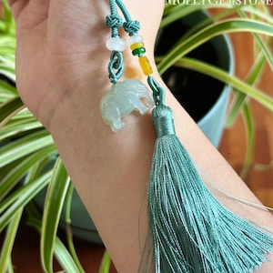 May include: A jade elephant pendant with a teal tassel and cord. The pendant is light green and translucent, with a detailed carving of an elephant. The cord features decorative knots and colorful beads. The text "Molly Gemstone" is visible in the background.