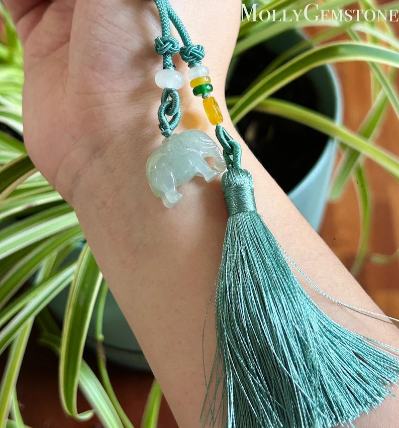 May include: A jade elephant pendant with a teal tassel and cord. The pendant is light green and translucent, with a detailed carving of an elephant. The cord features decorative knots and colorful beads. The text "Molly Gemstone" is visible in the background.
