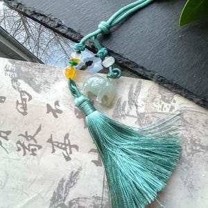 May include: A teal-colored tassel pendant with a small jade elephant charm, beads, and a knotted cord. The pendant is displayed on a paper with Asian calligraphy. The brand name "Molly Gemstone" is in the top left corner.