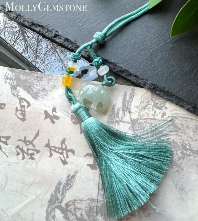 May include: A teal-colored tassel pendant with a small jade elephant charm, beads, and a knotted cord. The pendant is displayed on a paper with Asian calligraphy. The brand name "Molly Gemstone" is in the top left corner.