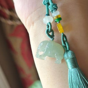 May include: A jade elephant pendant with a teal tassel and cord. The pendant is light green and has a detailed carving of an elephant. The cord has decorative knots and beads in white, yellow, and green.