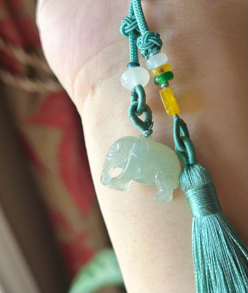 May include: A jade elephant pendant with a teal tassel and cord. The pendant is light green and has a detailed carving of an elephant. The cord has decorative knots and beads in white, yellow, and green.