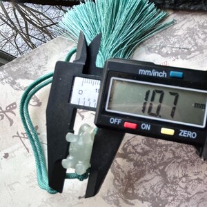 May include: A light green jade turtle pendant is measured with a digital caliper, showing a measurement of 10.7 mm. The pendant is attached to a green cord and a matching tassel. The background includes a patterned paper and a view of tree branches.