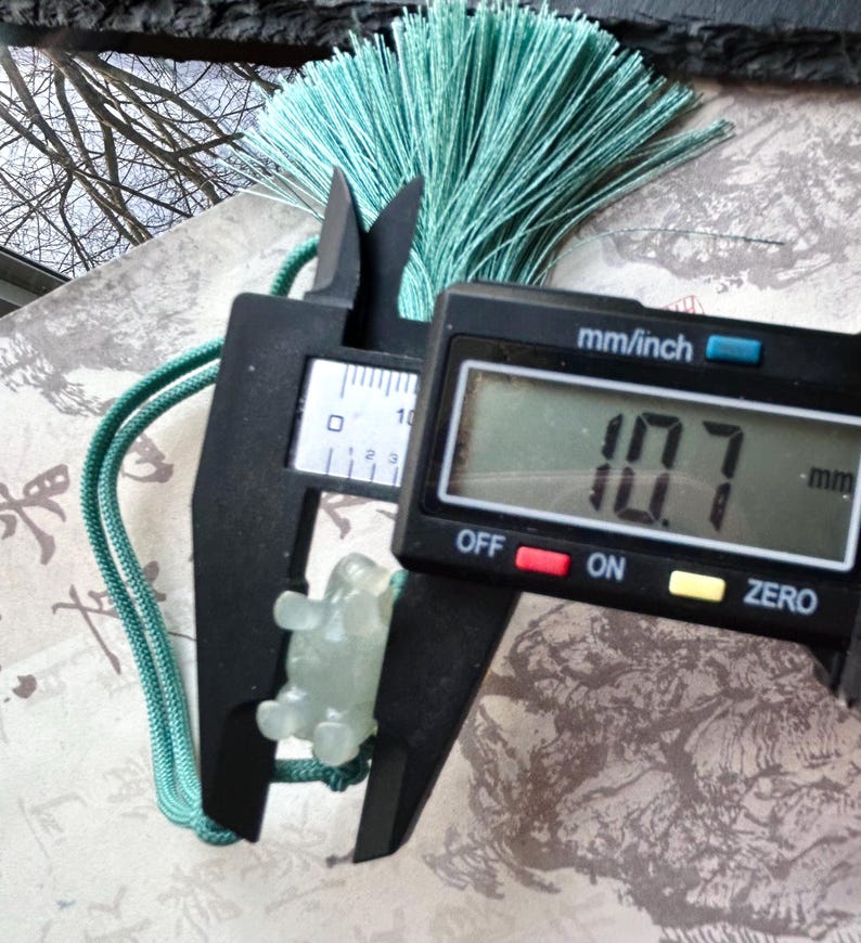 May include: A light green jade turtle pendant is measured with a digital caliper, showing a measurement of 10.7 mm. The pendant is attached to a green cord and a matching tassel. The background includes a patterned paper and a view of tree branches.