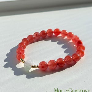 Könnte beinhalten: Ein rot-weißes Perlenarmband mit goldenen Akzenten. Das Armband besteht aus runden, durchscheinenden Perlen in Rot- und Weißtönen. Zwischen den roten Perlen befindet sich eine weiße, unregelmäßig geformte Perle. Das Armband ist mit dem Text "MollyGemstone" beschriftet.