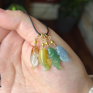 May include: A necklace featuring four carved stone fish pendants in translucent orange, yellow, green, and blue colors. Each fish is suspended from a gold-toned metal jump ring by a small gold bead and wire wrap, attached to a thin black cord.