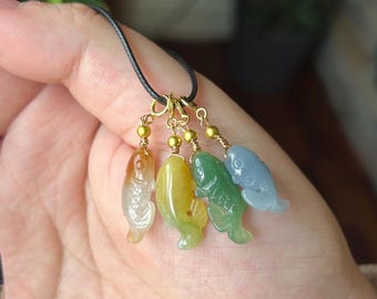 May include: A necklace featuring four carved stone fish pendants in translucent orange, yellow, green, and blue colors. Each fish is suspended from a gold-toned metal jump ring by a small gold bead and wire wrap, attached to a thin black cord.