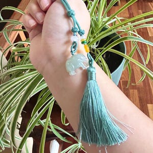 May include: A teal-colored elephant charm with a matching tassel. The charm is suspended from a teal cord with decorative beads. The elephant is carved from a translucent, pale green material.