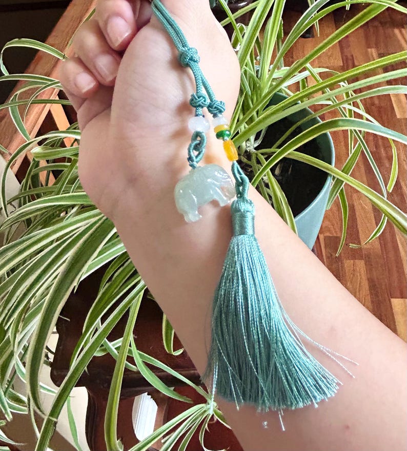 May include: A teal-colored elephant charm with a matching tassel. The charm is suspended from a teal cord with decorative beads. The elephant is carved from a translucent, pale green material.