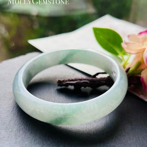 Puede incluir: Un brazalete de jade translúcido, con tonos verde pálido y blanco. La piedra pulida es circular y lisa. El brazalete se exhibe sobre una superficie oscura. El texto "MollyGemstone" es visible en la esquina superior izquierda.