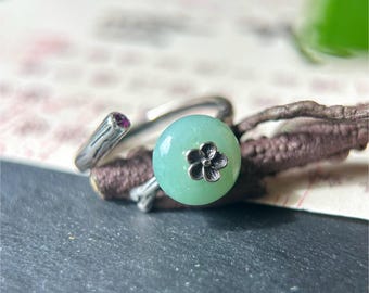 May include: A silver ring with a light green, circular gemstone and a small flower design. The ring has a branch-like design on the band and a small, purple gemstone. The text "MollyGemstone" is visible at the bottom.