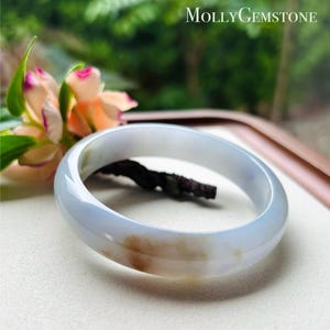 Op de afbeelding: Een lichtblauwe en bruine jade armband. Het gepolijste oppervlak van de armband reflecteert licht en toont de natuurlijke kleurvariaties. De armband heeft een cirkelvorm. Het Molly Gemstone-logo staat rechtsboven.