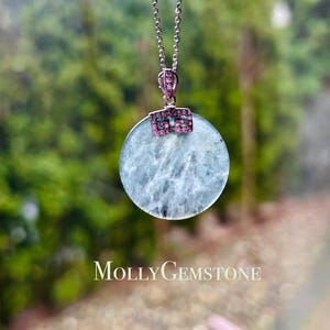 Könnte beinhalten: Eine silberne Kettenkette mit einem runden, hellblauen Anhänger mit einem silbernen und rosafarbenen Edelstein-Design. Der Anhänger ist von einem silbernen Rahmen umgeben. Die Kette ist vor einem verschwommenen grünen Hintergrund fotografiert.