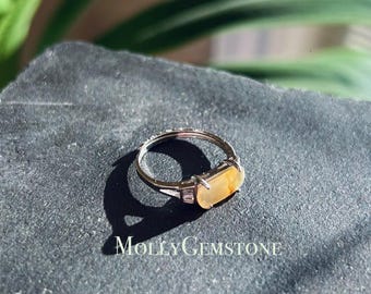 May include: A silver ring with a rectangular, light yellow gemstone set in the center. The gemstone is surrounded by small, clear gemstones. The ring is on a dark gray surface.