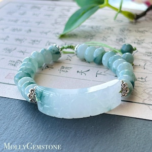 Könnte beinhalten: Ein Armband aus hellgrünem und weißem Jade. Es hat ein geschnitztes Jade-Mittelstück und runde Jade-Perlen. Silberfarbene Metalldetails akzentuieren das Armband. Der Text "MollyGemstone" ist unten sichtbar.