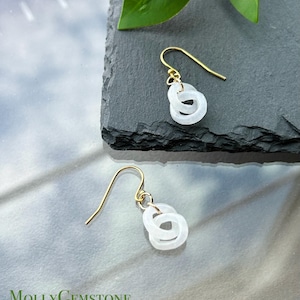 May include: A pair of gold-colored hook earrings with a white, interlocking circle design. The earrings are displayed on a dark gray surface with a green leaf in the background. The brand name "MollyGemstone" is visible at the bottom.