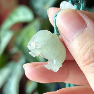 May include: A light green jade elephant pendant with a detailed carved design. The elephant is suspended from a teal green cord with a small white bead. The pendant is held in a person's hand.