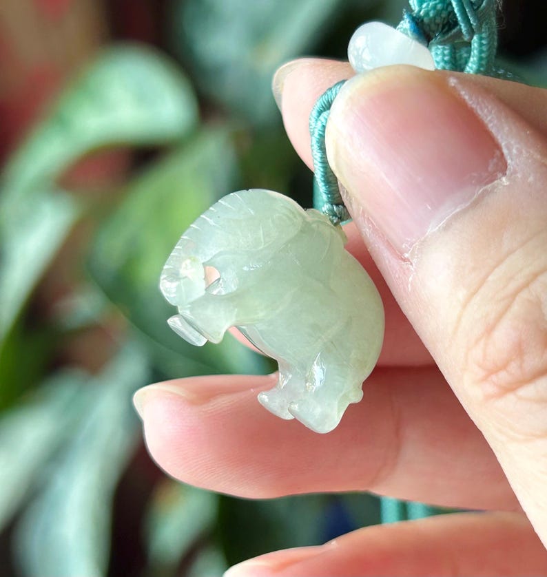 May include: A light green jade elephant pendant with a detailed carved design. The elephant is suspended from a teal green cord with a small white bead. The pendant is held in a person's hand.