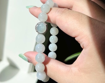 May include: A jade bracelet with a round, light green jade bead in the center. The bead is surrounded by a silver setting with small diamonds. The bracelet is made of round, light green jade beads.