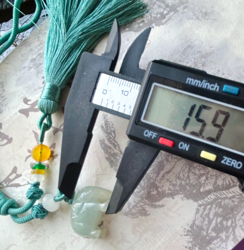 May include: A jade elephant pendant with a teal tassel and beaded accents. The pendant is being measured with a digital caliper, showing a width of 15.9 mm. The pendant is a light green color.
