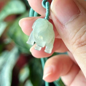 May include: A light green jade elephant pendant on a matching cord. The elephant is carved with visible legs, trunk, and ears. The pendant is held up by a green cord, and the background is blurred.