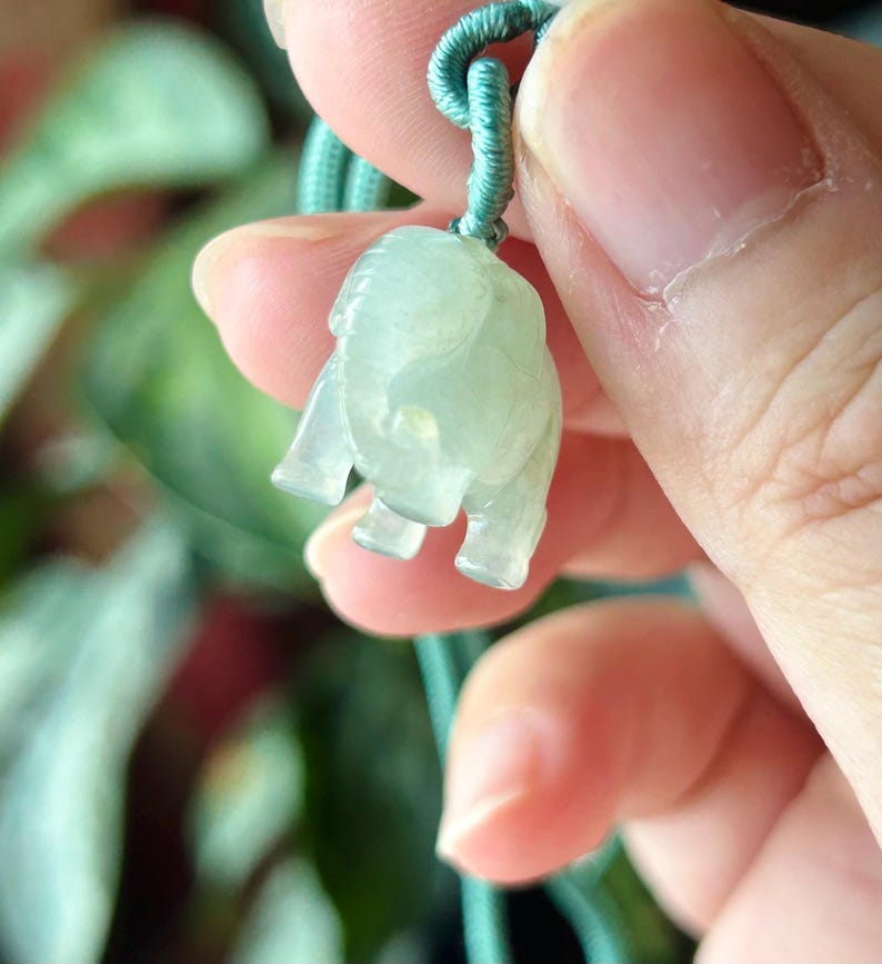 May include: A light green jade elephant pendant on a matching cord. The elephant is carved with visible legs, trunk, and ears. The pendant is held up by a green cord, and the background is blurred.