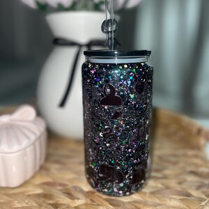 Peut inclure: Un gobelet noir &agrave; paillettes avec un couvercle noir et une paille transparente. Le gobelet est d&eacute;cor&eacute; de divers motifs de silhouettes de couleur fonc&eacute;e. Le gobelet est pos&eacute; sur une surface tiss&eacute;e, avec un vase blanc et des fleurs roses en arri&egrave;re-plan.