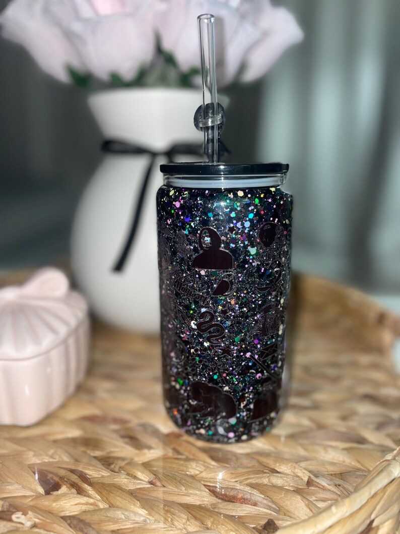 Peut inclure: Un gobelet noir &agrave; paillettes avec un couvercle noir et une paille transparente. Le gobelet est d&eacute;cor&eacute; de divers motifs de silhouettes de couleur fonc&eacute;e. Le gobelet est pos&eacute; sur une surface tiss&eacute;e, avec un vase blanc et des fleurs roses en arri&egrave;re-plan.