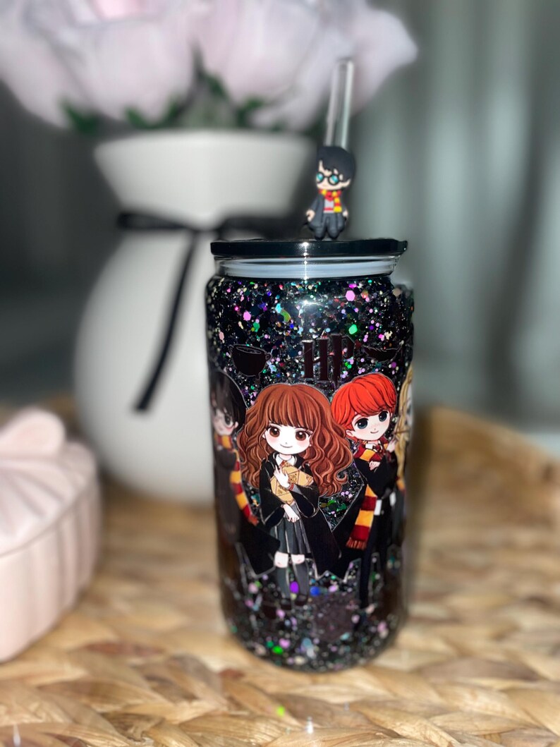 Peut inclure: Un gobelet noir &agrave; paillettes avec des personnages de dessins anim&eacute;s d'une s&eacute;rie fantastique populaire. Le gobelet a un couvercle noir, une paille transparente et une petite figurine sur le dessus. Les personnages sont en uniforme scolaire.