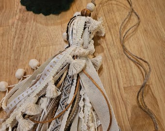 May include: A handmade tassel with various fabric textures and colors, including cream, beige, and gold. The tassel features decorative ribbons, tassels, and wooden beads. The words "FAITH" and "LOVE" are engraved on the beads. A lit candle in a glass cup is in the background.