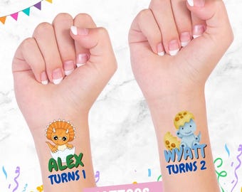 May include: Two temporary tattoos, one with a blue dinosaur and the text "WYATT TURNS 2" and the other with an orange dinosaur and the text "ALEX TURNS 1".
