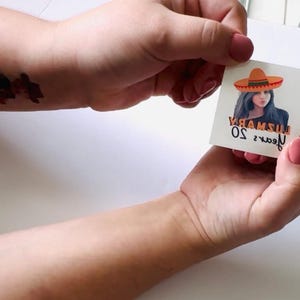 May include: A temporary tattoo depicting a woman in a sombrero. The image includes the text "YAMSU 50 years". The tattoo is held between two hands with pink nail varnish. A puzzle piece tattoo is visible on the wrist.