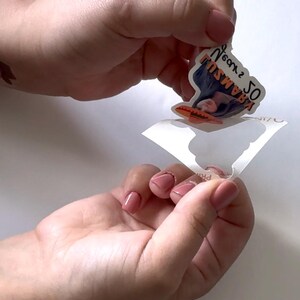 May include: A sticker featuring a woman's face and the text "50 KRAMSU" in orange and black. The sticker is being peeled from its backing, revealing a white surface.