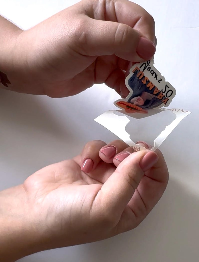 May include: A sticker featuring a woman's face and the text "50 KRAMSU" in orange and black. The sticker is being peeled from its backing, revealing a white surface.
