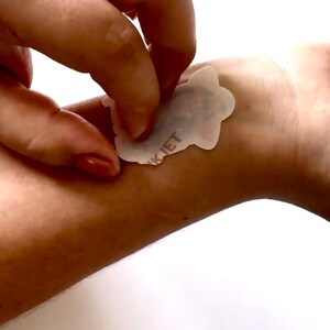 May include: A person is peeling a small, white, die-cut sticker from their wrist. The sticker has a black design and the word "K JET" printed on it. The skin tone is light, and the background is a plain white.