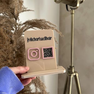 May include: A beige acrylic display stand with a pink Instagram logo and a QR code. The text "kylichartwellhair" is printed in black. The stand is held in front of a dried plant and a brass-coloured spotlight.