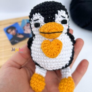 Pode incluir: Um pinguim de crochê feito à mão, com cabeça e costas pretas, barriga branca e bico, pés e coração laranja. O pinguim tem os olhos fechados e é segurado em uma mão. O logotipo "Jo ba" é visível no fundo.