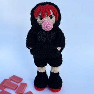 Pode incluir: Boneca de crochê feita à mão usando um moletom com capuz e shorts pretos. A boneca tem cabelo ruivo, uma bolha de chiclete rosa e sapatos pretos com solas vermelhas. A boneca está em pé.