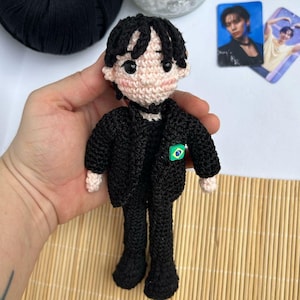 Pode incluir: Boneca de crochê vestindo um terno preto com uma pequena bandeira verde e azul no peito. A boneca tem cabelo preto e rosto rosa claro.