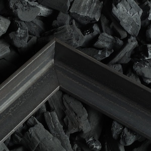 Könnte beinhalten: Zwei schwarze Metallecken, möglicherweise für einen Rahmen, liegen auf einem Bett aus schwarzen Holzkohlebriketts.