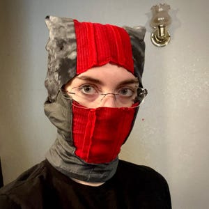May include: A handmade balaclava featuring a patchwork design. The head covering combines gray and black patterned fabric with a vibrant red panel across the forehead and face. The neck portion is gray.