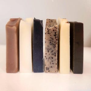 May include: A collection of six rectangular bars of handmade soap in various colors. The colors include shades of brown, white, black, speckled tan, cream, and dark green. The soaps are arranged vertically, side by side.