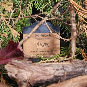 May include: A bar of dark gray soap with a brown paper label that reads "DK Naturals Tallow Soap Hunter Unscented for Hunting Purposes Net Wt. 3.5oz-4oz". The soap is surrounded by green branches.